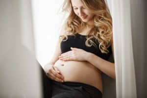 close-up-detail-young-blonde-pregnant-mother-black-outfit-sitting-window-sill-bedroom-touching-looking-belly-with-happy-expression
